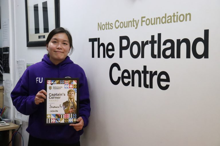 Stephanie Fu in her branded purple hoodie, stand proud with her Captain's Corner certificate next to the Portland Centre sign in the reception area.