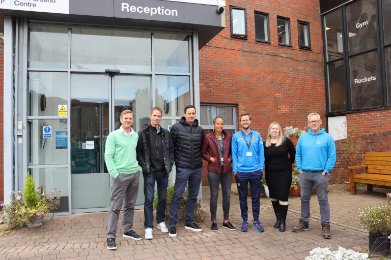 The image shows a group of seven individuals standing outside the entrance of the Portland Centre. The group includes men and women dressed casually and in sportswear, posing together in front of the building's glass doors with a "Reception" sign above. The brick exterior and windows display the words "Gym" and "Rackets" indicating the centre’s facilities. There are potted plants and flowers on either side of the entrance, adding a welcoming touch to the scene.