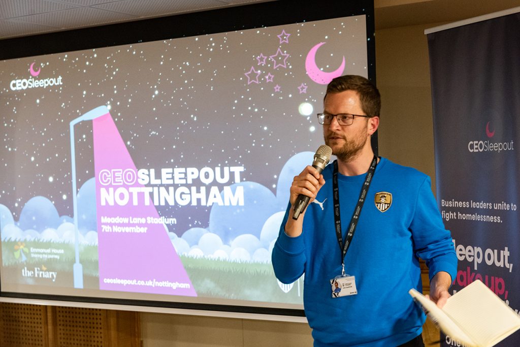 Sam Crawford of Notts County Foundation is wearing a blue sweatshirt with the Notts County FC logo and holding a microphone and speaking in front of a presentation screen. The screen displays the title 'CEO Sleepout Nottingham' with event details, including 'Meadow Lane Stadium' and the date '7th November.' The background features an illustrated night sky with stars, a crescent moon, and clouds. To the right, a banner reads, 'Business leaders unite to fight homelessness.