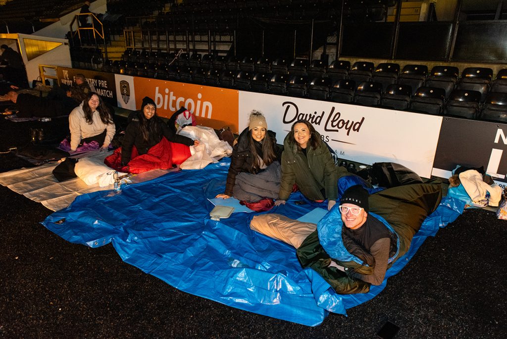 "A group of participants sitting on a large blue tarp at the CEO Sleepout and sleeping bags. They are smiling. Behind them are stadium seats and advertising boards for businesses, including 'David Lloyd Clubs' and 'Bitcoin.'"